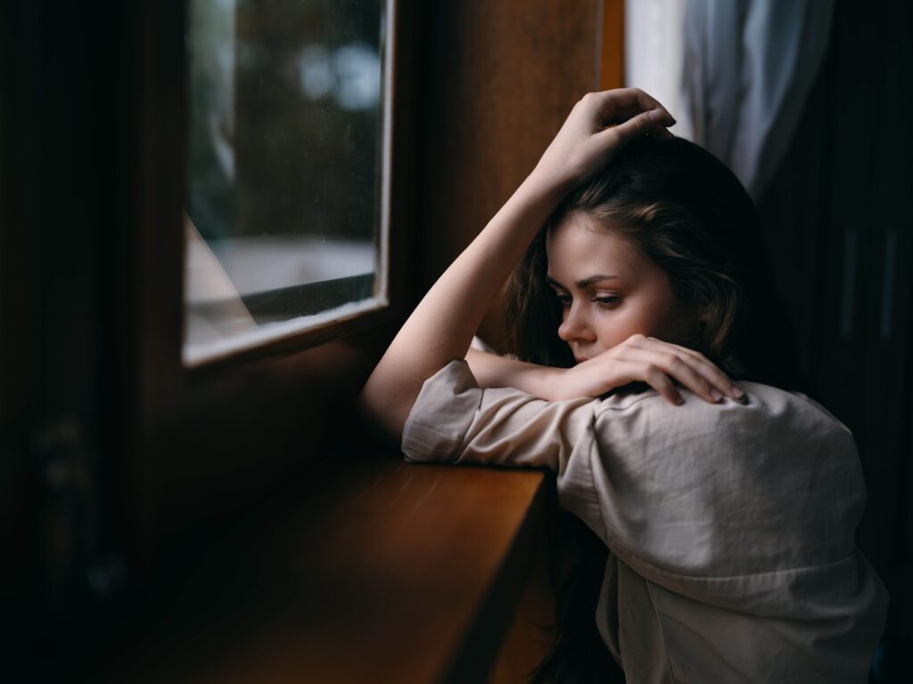 Sad woman by window, struggling with C-PTSD.