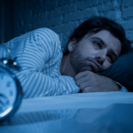 A man lying in bed, looking at an alarm clock, illustrating signs of insomnia and sleep difficulties.