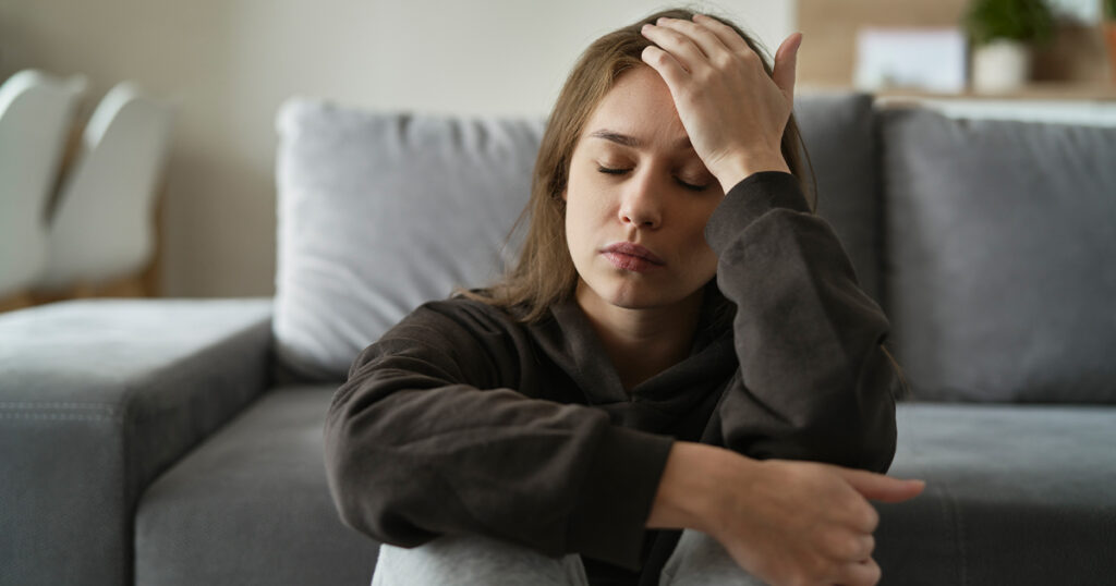 A woman sitting on a couch, looking distressed with her hands on her head, reflecting feelings of guilt and frustration.