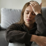 A woman sitting on a couch, looking distressed with her hands on her head, reflecting feelings of guilt and frustration.
